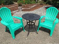 Two teal plastic Adirondack chairs with a black round metal table between them, positioned outdoors on a paved surface.