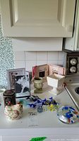 Photo showing glass candies, Murano marked paperweight, frames, tin, mini planters, and basket arranged on kitchen counter.