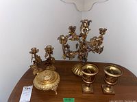 Overview photo showing all items together on wooden surface against white wall corner. Includes large candelabra, smaller double candle holders, lidded container, and miniature urns.