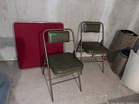 Two folding metal frame chairs with olive vinyl seat and back next to a folded red padded card table.