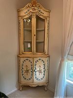 Full view of cream painted corner cabinet in a room corner showing upper glass doors, lower painted doors, carved crown and legs.