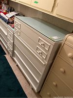 White painted dresser with five drawers, ceramic floral knob pulls, clear glass top with chips along edges. Some marks on paint. Positioned indoors next to other furniture.