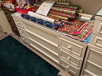Front view of white painted wood dresser with six side drawers, three middle drawers, ceramic floral drawer pulls, and clear glass top. Wrapped gift paper on top.