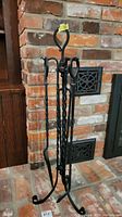 Full view of the iron fireplace tool stand holding tools, set against a brick background
