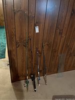 Full view of the four fishing rods standing against a wood wall showing two rods with reels and two without.
