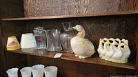 Shelf with two glass vases, clear glass jar with lid, Fitz and Floyd goose cookie jar, geese pottery piece, and small fruit-themed lamp shades.