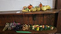 Overview of all fruit decorations on wooden shelf including gourds, pumpkins, bananas, glass grapes, plastic grapes, and wooden fruit wall hangings below.