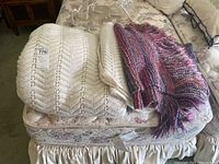 Photo shows three folded knit blankets stacked on a bed with floral bedding, featuring a large off-white blanket, a smaller cream blanket, and a colorful fringed throw.