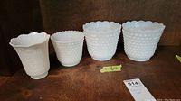 Four white milk glass hobnail vases on wooden shelf, including one square-based and three round-based with scalloped edges.