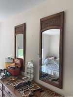 Two vertical rectangular wall mirrors with wood frames featuring decorative leaf accents above the reflective surface. Mirrors are shown hanging on a beige wall above a dresser with jewelry and various items.