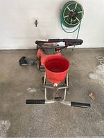 Wide view of the red lawn mower with attached side grass catcher and front handlebar, and red plastic bucket fertilizer spreader mounted on metal frame with dual black handles in front.