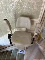 Front-facing view of beige padded seat and armrests on stair lift, installed on rail attached to staircase.