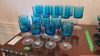 Seventeen blue glass wine glasses displayed on a wooden surface, showing both shorter and taller sizes clearly.
