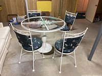 Photo showing round glass-top cafe table with white metal pedestal base and four matching chairs with floral upholstery.