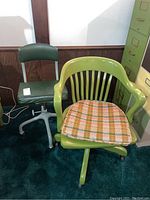 Two vintage office chairs next to each other, one green leather swivel chair with metal frame and casters, and one green wooden swivel chair with plaid cushion.