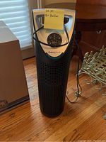 Thera Pure tower air purifier standing on wooden floor near chair and box. Shows front digital display with control buttons and attached power cord.