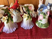 Five porcelain bird bells arranged on a red fabric surface including three with green hummingbirds on purple skirt bases with plumeria flowers, one with a red cardinal and holly-accented white base, and one with a blue bird and floral-decorated base.