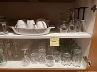 Front view of upper and lower cabinet shelves showing white dishes, serving dish, clear stemmed wine glasses, and various tumblers.