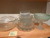 Multiple clear glass bowls with different sizes and shapes, including a frosted footed bowl with scalloped edge, stacked in a shelf.