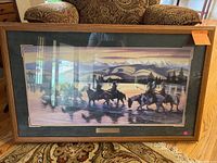 Framed western print showing cowboys herding horses near water with mountains in background. Dark green frame with glass.