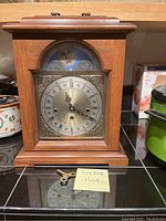 Front view of the wooden mantel clock with brass-tone Roman numeral dial and moon phase feature. Clock sits on counter with metal winding key in front.