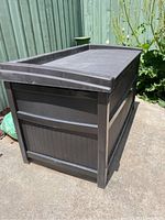 Black plastic outdoor storage bench against a green fence on a concrete surface, dusty condition, showing side and corner view.