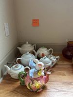 Six assorted tea pots displayed on a wooden countertop including a white teapot, floral designs, and a bunny-shaped teapot. The photo shows overall lot contents.