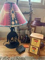Western-themed lamp with fabric shade, small drawers with southwestern design, rustic ladder, metal horse decoration, and double cone lamp with cup holders placed on wood floor and against wall under window.