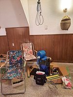 Wide shot showing two foldable camping chairs with floral upholstery, blue plastic water dispenser, camping gear boxes, and folding stools.