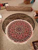Two round carpets placed on beige carpeted floor, smaller red-centered carpet on top of larger darker carpet, fringes visible on smaller rug, rolled carpet in background.