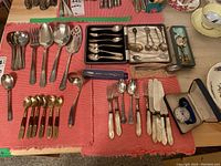 Wide view of mixed flatware and utensils laid on red cloth showing forks, knives, spoons, and a boxed Gallet stopwatch.