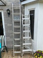 Two aluminum ladders side by side, one smaller folding ladder and one larger extendable ladder, both standing upright outdoors.