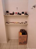 Shelf with assorted camera lenses, filters, lens caps, and Kodak Carousel slide trays in box on floor below.