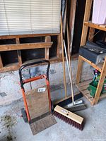 Movers dolly with three push brooms leaning against a garage wall near window and shelving.