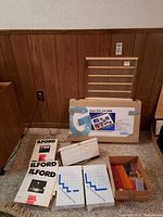 Photo of boxed Gepa Slim Lite 5000 slide viewer, Ilford photographic paper packs, transparent slide lightbox viewer and various slide viewer accessories on carpet against wood panel wall.