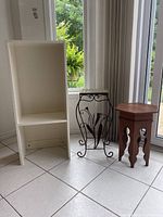 Photo showing the three items on tile floor near windows: a white laminated shelf, black wire frame plant stand with decorative metal flowers, and a small wooden plant stand.