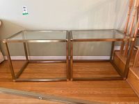 Front view of two metal and glass side tables side by side on wooden floor against wall, showing clear glass tops and brushed metal frames.