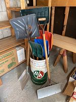 Photo showing a group of snow shovels resting inside a vintage British American Oil Company drum in a garage environment.