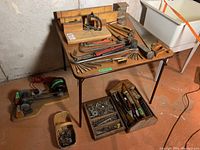 Photo showing an overview of vintage tools including hammers, crowbars, saw, and hardware trays on a table in a basement setting.