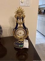 Full view of blue and gold lyre-shaped mantel clock with sunburst female face ornament on top, and Austrian crystals trim around clock face.
