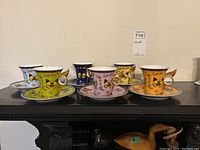 Full set of six coffee and tea cups with matching saucers in different colors and butterfly designs arranged on a shelf.