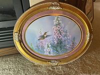 Oval framed print of a Rufous Hummingbird flying by pink foxglove flowers, framed in gold ornate frame with pink matte and gold trim.