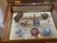 Top view of all items displayed in glass cabinet including Wedgwood lidded dish, paperweights, Royal Crown Derby bird, Spode plate, Ayshford teapot, and oriental music box