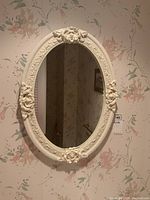 Full front view of the oval mirror hanging on a floral wallpapered wall showing overall shape and decorative frame.