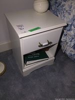 Front view of one white night stand showing drawer with brass and white handle and open lower shelf containing books.