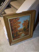 Framed oil painting leaning against a carpeted stair with autumn forest scene and path.