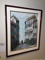 Framed painting showing a street with flower-decorated buildings and a person walking, viewed from an angle