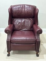 Front view of burgundy leather reclining chair showing padded back, arms with brass studs, and carved wood claw and ball feet.
