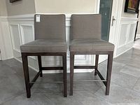Pair of grey fabric bar stools front view showing matching cushions and dark wooden legs.