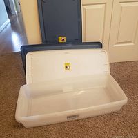 Open empty clear rectangular plastic under bed storage box with white lid in background and two blue lids stacked behind.